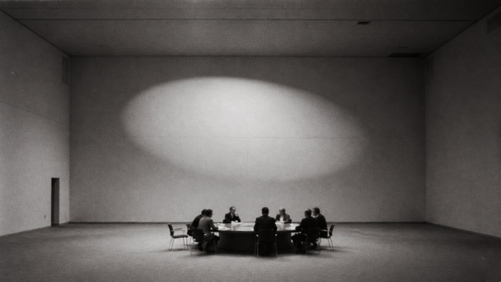 A small group seated around a round table in a vast, dimly lit room.